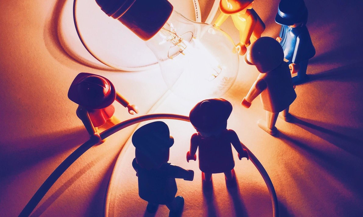 Toy people in a group around a light bulb. The bulb illuminates the picture and gives them shadows to say people are enjoying the theme of gamification and games.