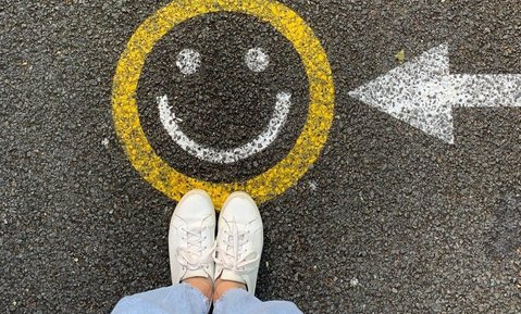 A smiley drawn on the road. A person's feet in white canvas shoes is shown below it. the the right, there is an arrow pointing towards the face.
