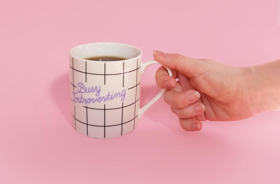 A coffee mug with the words busy introverting in a pink background