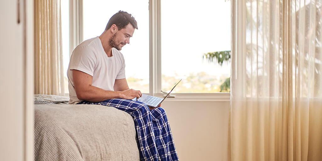 photo of a man working on a laptop, sitting on the edge of his bed in his pyjamas
