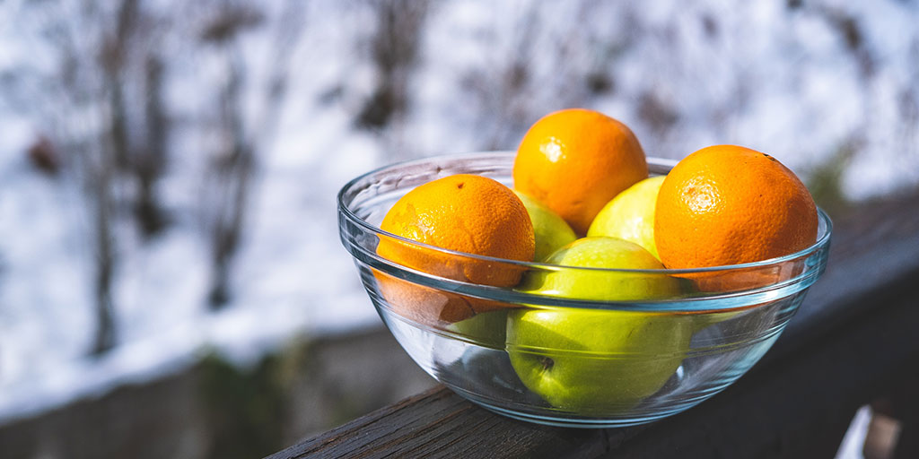 image of some apples and oranges in a glass bowl on a wooden ledge with some snow in the background